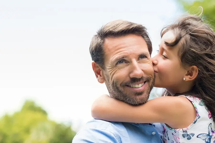 Smiling father being hugged and kissed on the cheek by his daughter outdoors, reflecting restored family bonds in recovery.