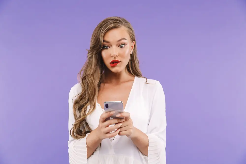 Woman in white blouse looking surprised at her smartphone against a purple background.