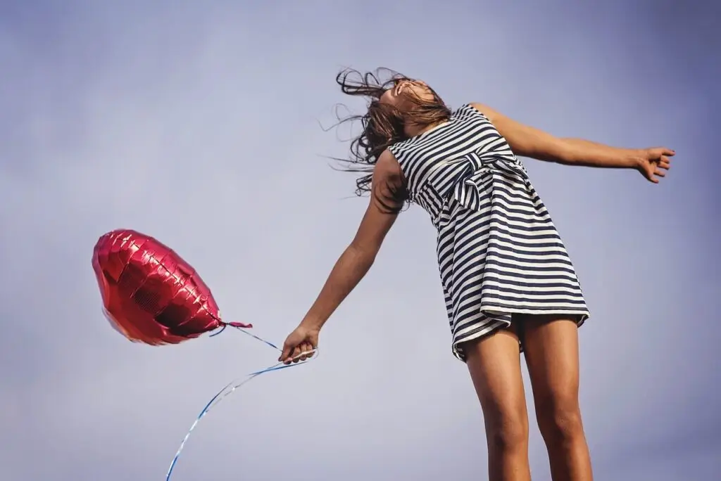Woman in striped dress holding a red heart-shaped balloon, leaning back joyfully against a blue sky.
