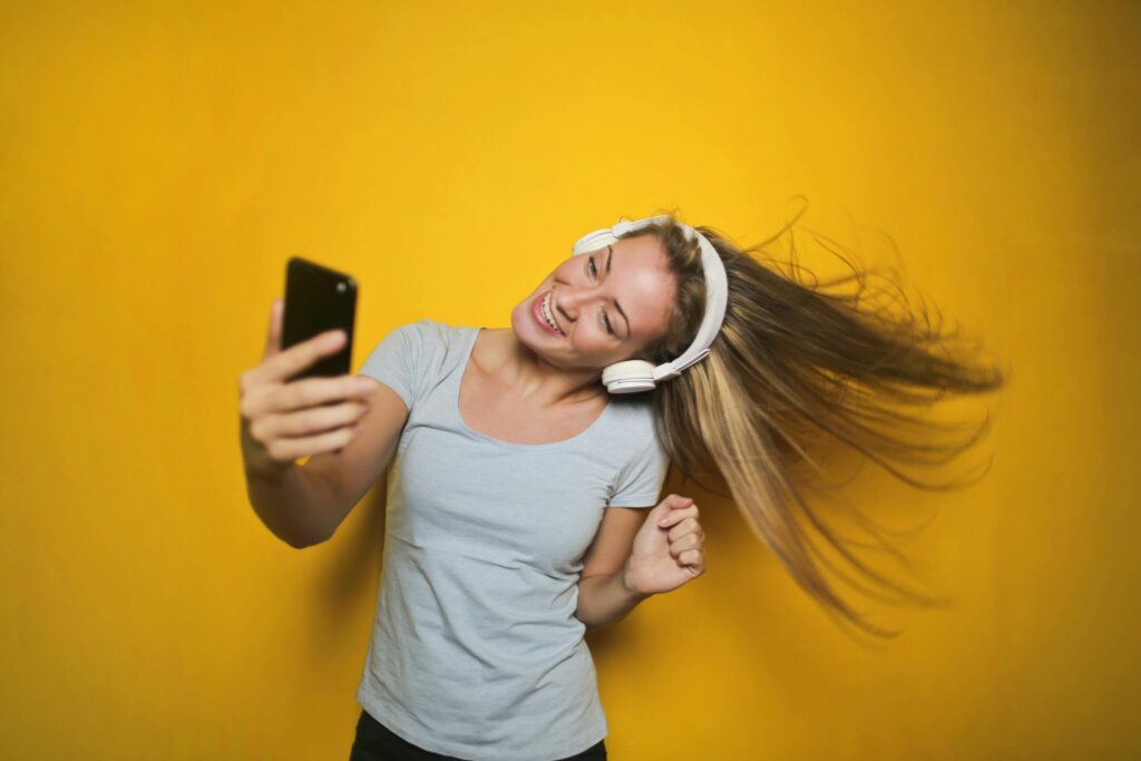 Smiling woman with headphones dances and takes a selfie against a bright yellow background.