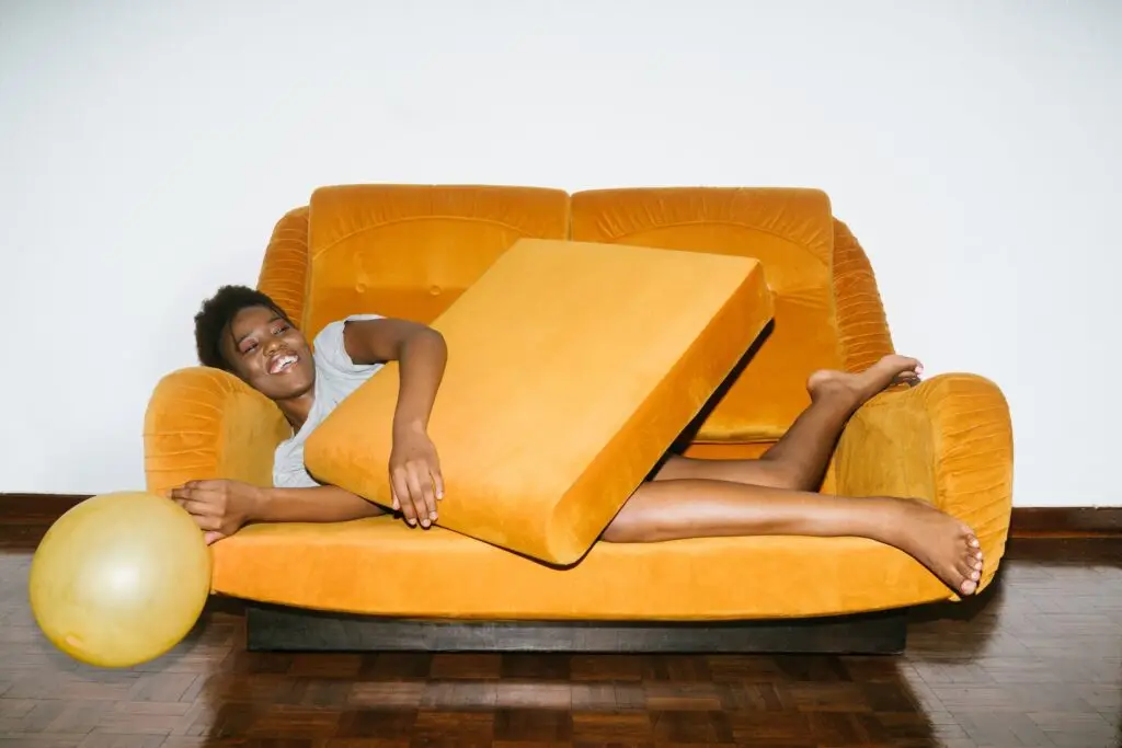 Smiling woman lying on a yellow couch holding a cushion, with a balloon on the wood floor beside her.